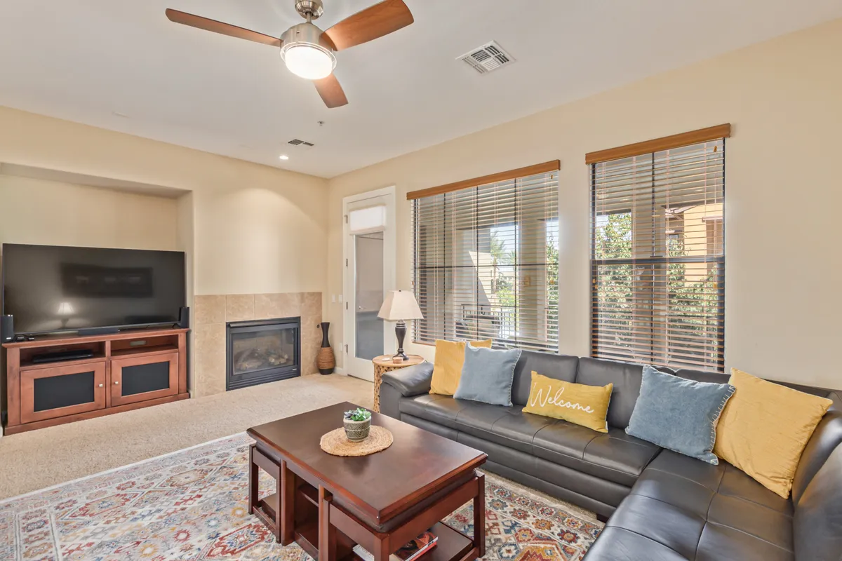 A cozy living room filled with natural light, showcasing a comfortable sofa with decorative pillows, a sleek coffee table, a smart television mounted above a modern fireplace, and large windows with blinds that flood the space with brightness. The area is set up for relaxing, entertaining, or watching TV, with a warm, inviting atmosphere.
