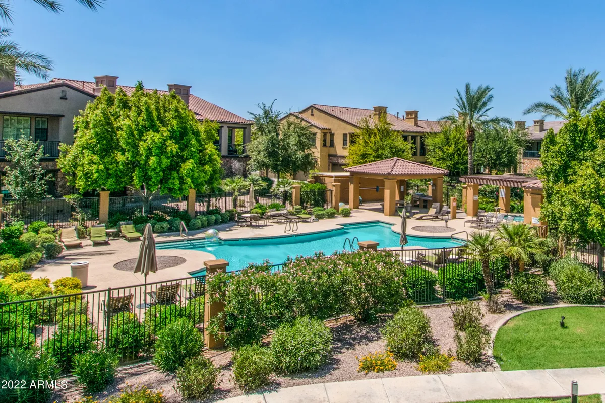 A sparkling swimming pool sits at the heart of the community, surrounded by lush greenery and an inviting outdoor seating area. The backdrop includes the surrounding residential buildings, creating a sense of a close-knit, vacation-style neighborhood. The tranquil blue water, ample seating, and manicured landscape evoke relaxation and togetherness.