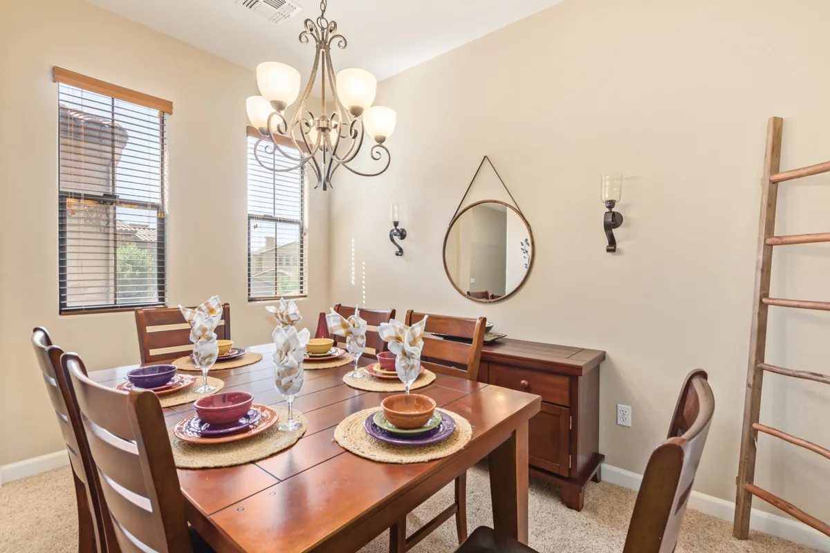 A sunlit dining area with a table that comfortably seats six, positioned under a stylish chandelier. The mirrored wall, window with blinds, and inviting table setting evoke a sense of warmth, making it ideal for family meals or entertaining guests.