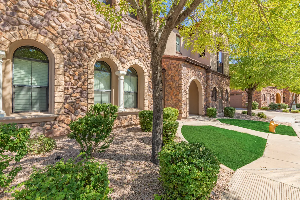 A charming exterior view of Serenity Town House’s stone facade, accentuated by arched windows and surrounded by lush greenery. The pathway leading through the landscape is ideal for morning strolls, and neighbors can be seen enjoying the shade under mature trees.