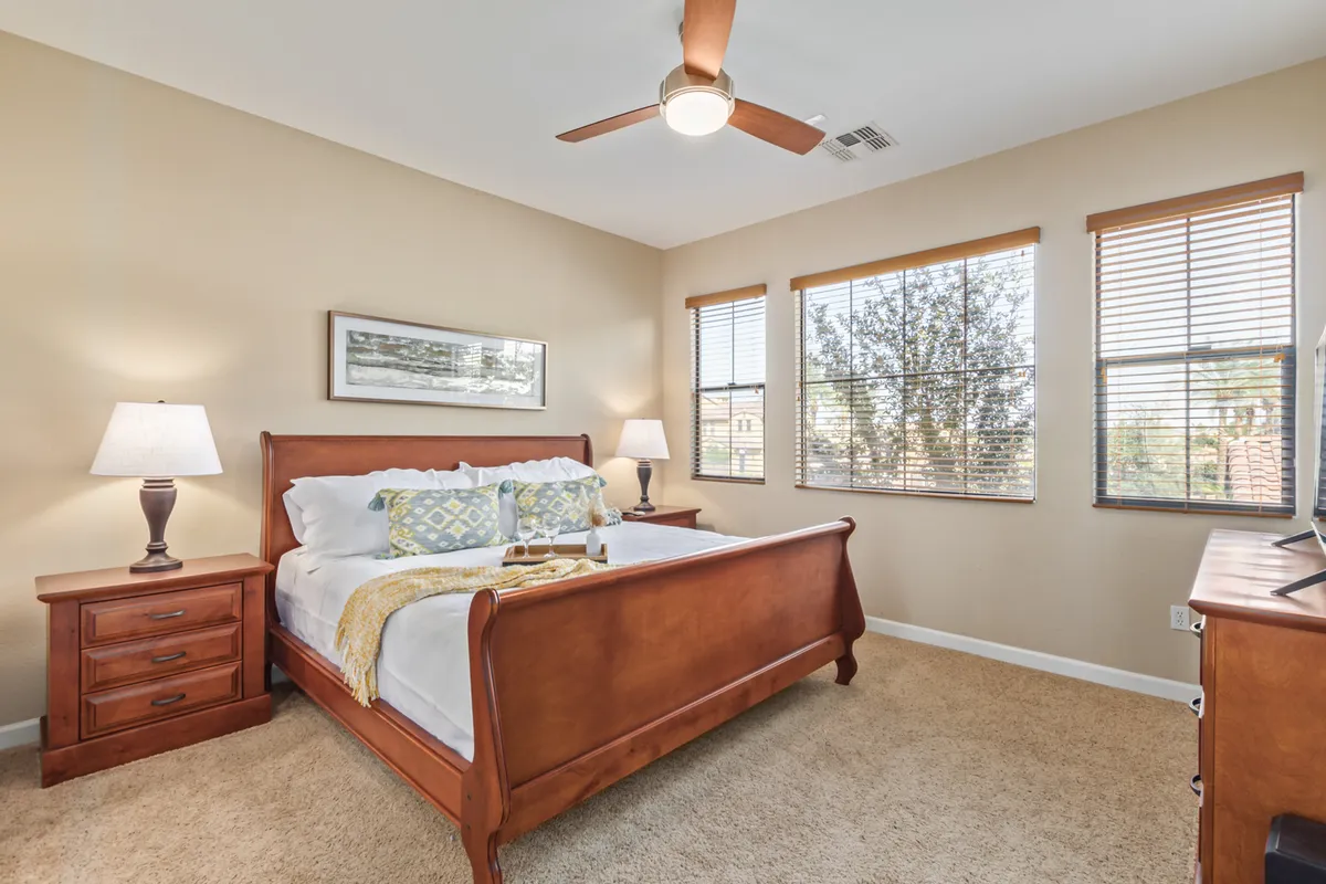 A spacious bedroom featuring a comfortable bed flanked by nightstands, large windows letting in natural light, and a ceiling fan above. The room exudes a sense of calm with its uncluttered decor and soft, inviting linens, perfect for rest and relaxation.