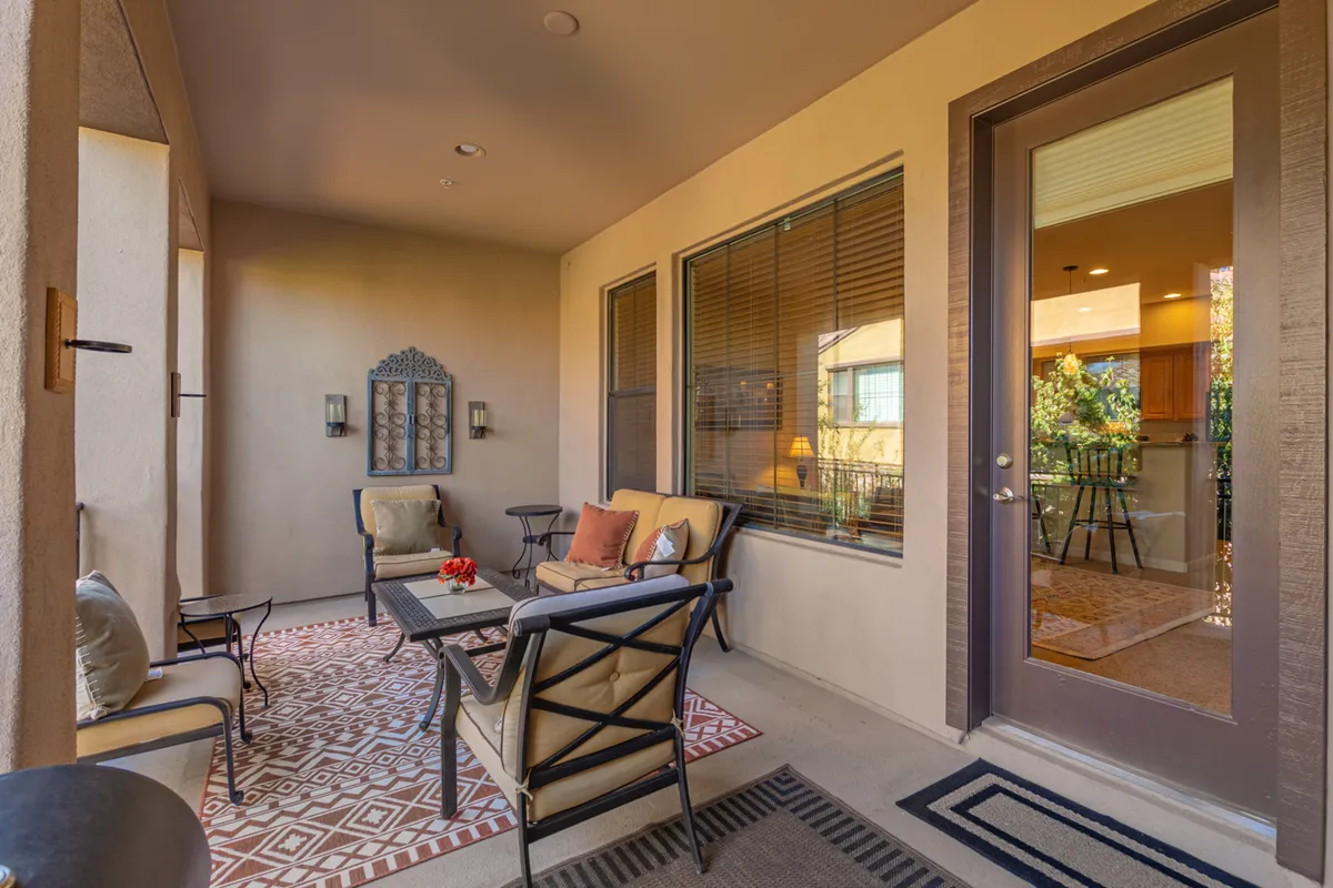 A welcoming and stylish outdoor seating area on a spacious balcony, featuring comfortable chairs, a decorative wall mirror, and a textured area rug. The area is bathed in warm light, inviting relaxation and social gatherings. You can see lush landscaping and sunset views in the distance, enhancing the sense of a private retreat.