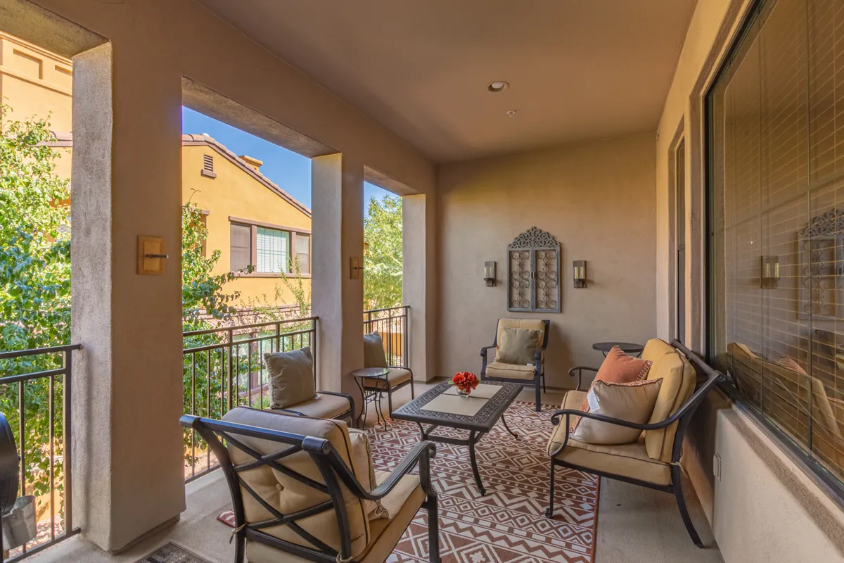 A serene exterior seating area with comfortable outdoor furniture, decorative wall art, and a cozy textured rug. Gentle sunshine filters through trees, casting a pleasant glow over the space. The area is ideal for socializing, sipping coffee, or enjoying tranquil views of the landscaped grounds.