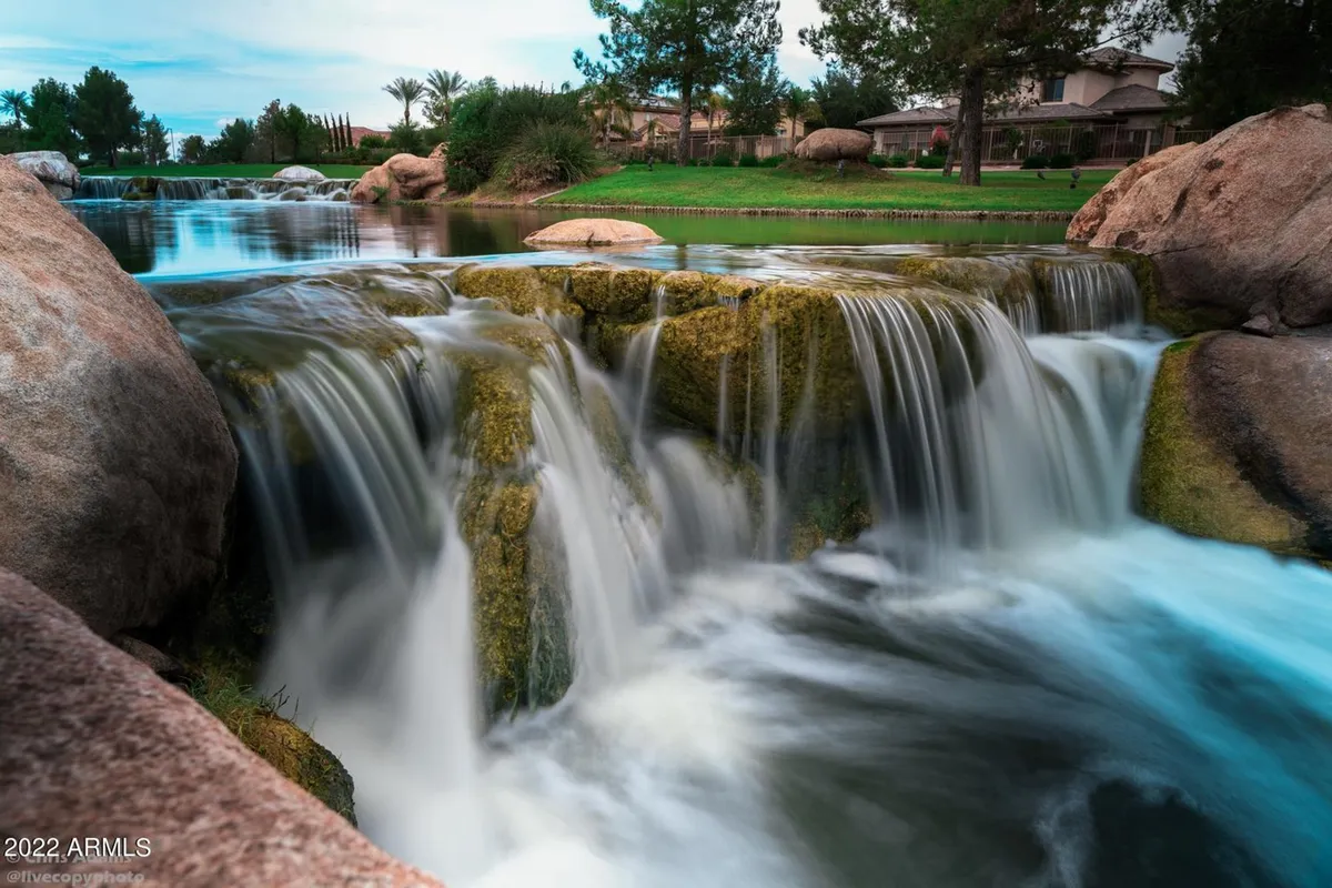 A serene natural setting with a picturesque waterfall flowing over smooth rocks into a reflective pool, surrounded by lush greenery. The area is ideal for relaxing by the water, picnicking, fishing, or enjoying nature walks.