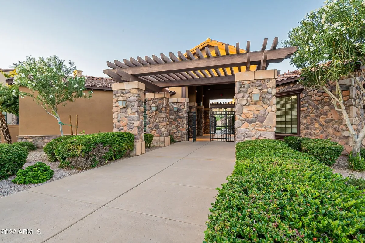 A warm exterior shot of the townhouse entrance, featuring a stone pathway, a wooden pergola, and lush green shrubs. The inviting setting suggests a safe, quiet, and family-friendly environment within a gated community.
