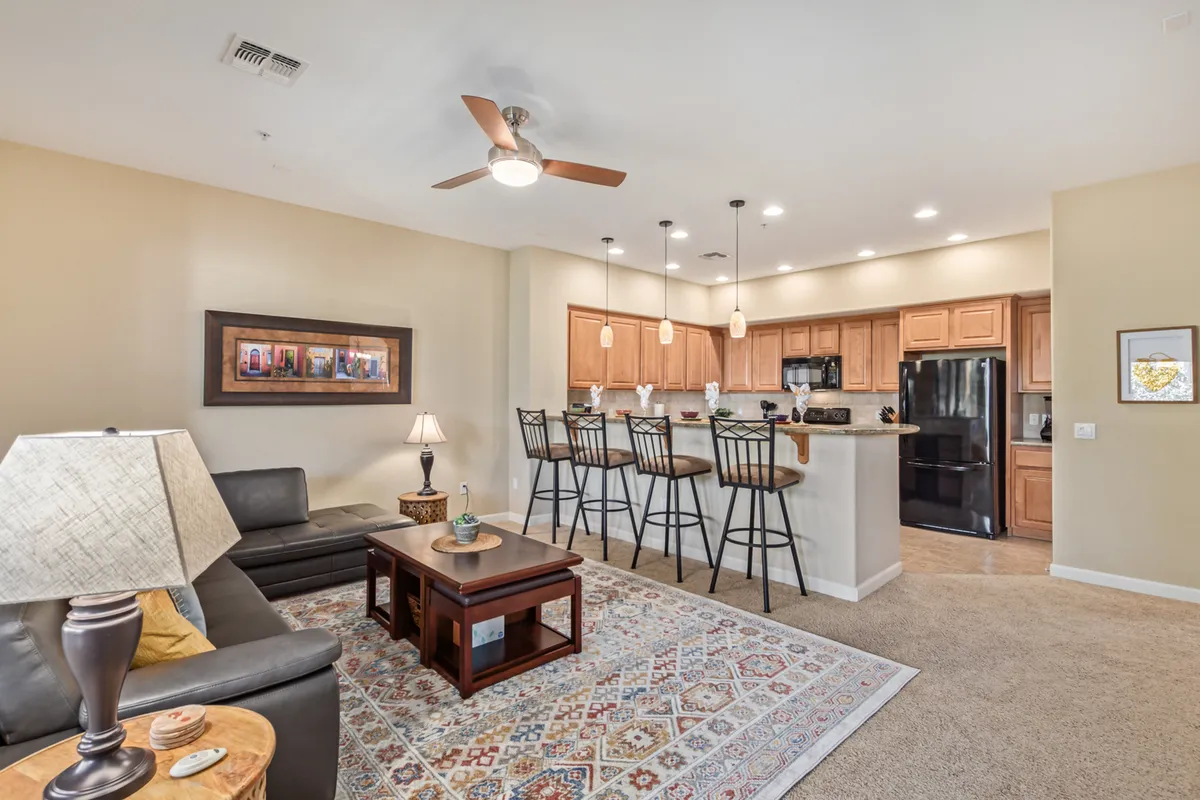 An open sightline from the living area to the kitchen, featuring a sofa and coffee table in the foreground, a kitchen island with bar stools, and cheerful artwork. The space is unified by a neutral palette, creating a seamless transition between lounging and meal prep areas.